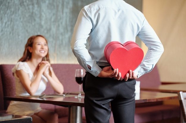 Man hiding romantic present for girlfriend behind his back when standing at restaurant table