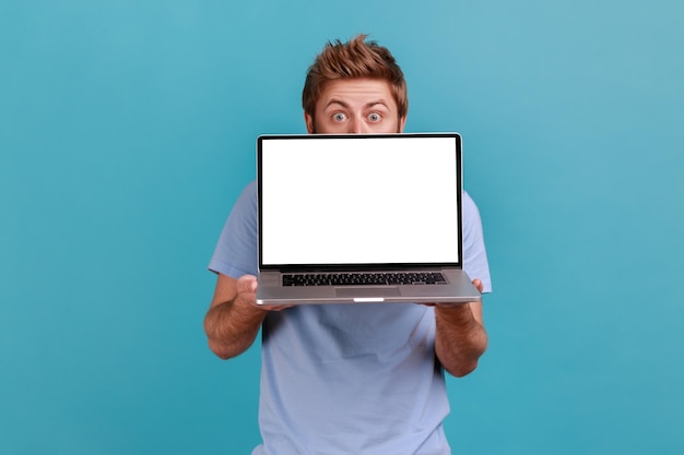 Man hiding behind white screen laptop surprised with advertising area looks at camera with big eyes