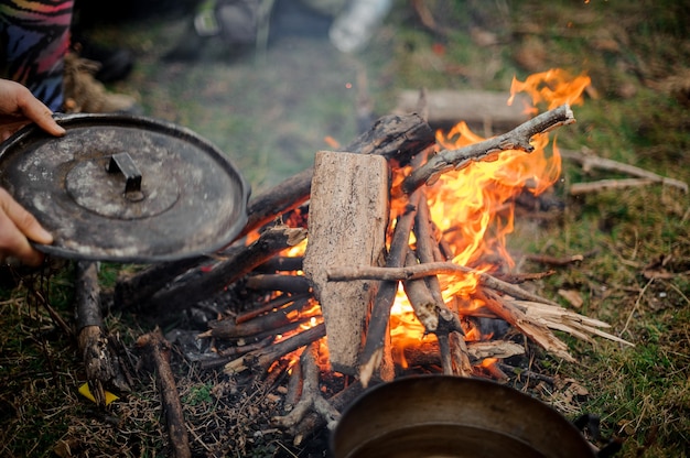 Man holding cover placing on the pan near the campfire with wood in the green forest