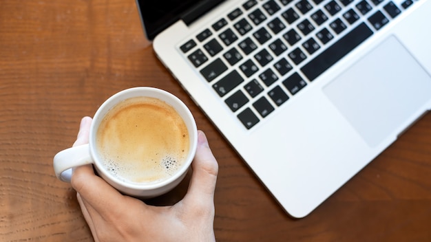 A man holding a cup of coffee, laptop on the wooden table. Top view