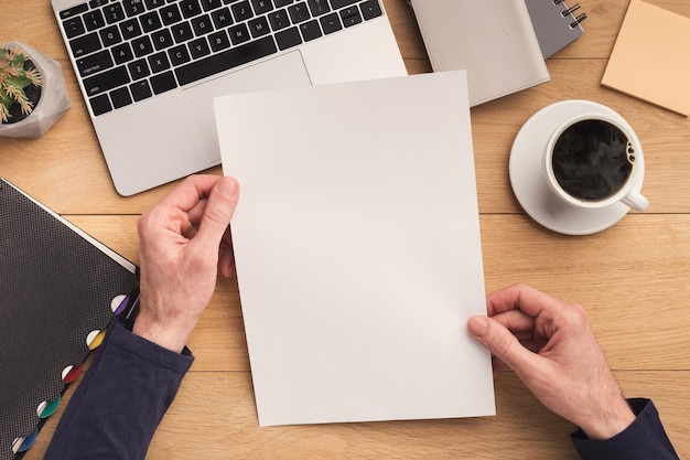 Man holding empty sheet of paper for advertisement and drinking coffee on vintage wooden table, top view. Male workplace with cacti and notebook