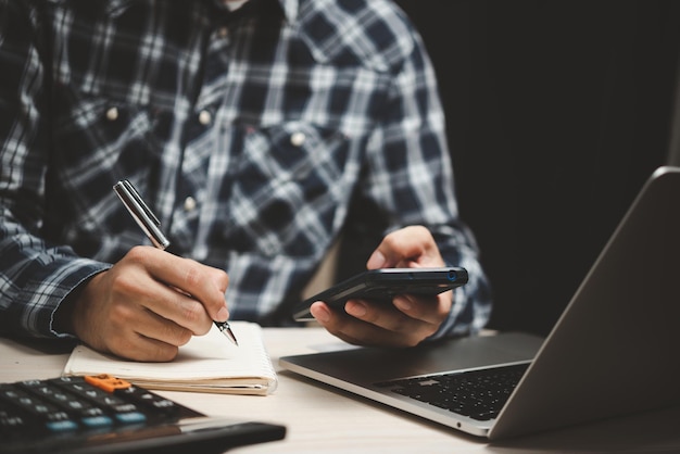 Man holding a phone with a pen and paper to write a note in an office diary using a laptop at office