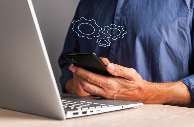 Man holding phone with virtual gears while working on laptop Mobile settings or strategy process organization business idea concept