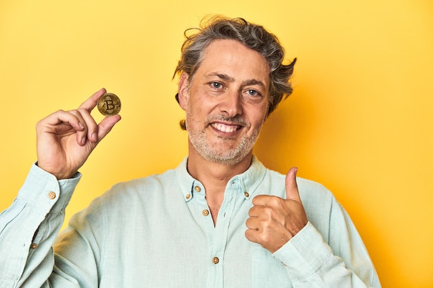 Man holding a physical bitcoin yellow backdrop smiling and raising thumb up