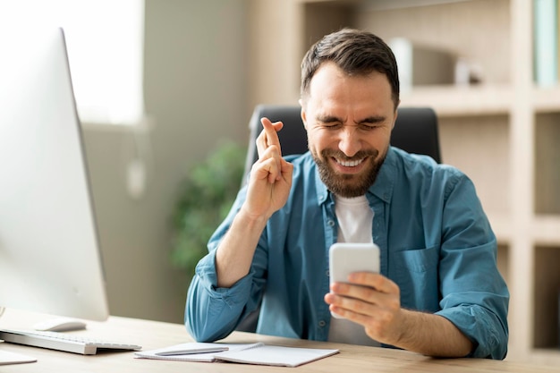 Man holding smartphone and keeping fingers crossed sitting at desk in office