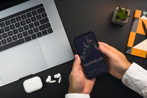 Man holding a smartphone with financial stock market graph on the screen on black background table Office environment Stock Exchange