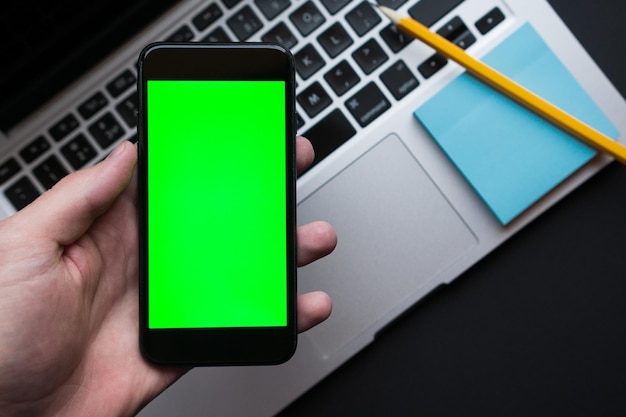 Man holding smartphone with green chroma key screen on background modern black office desk table with laptop Modern workplace