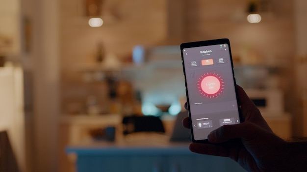 Man holding smartphone with lighting control application, turning on the lights sitting in kitchen house with automation lighting system. Person using smart home software working remote at laptop