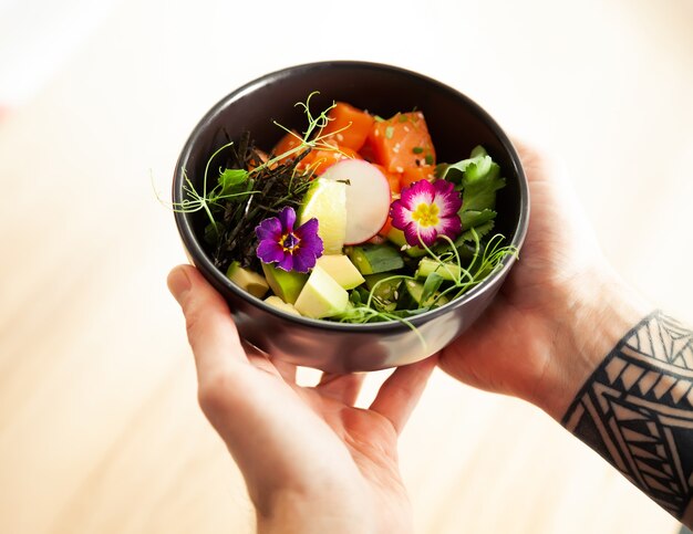 A man holds a bowl of poke salad with salmon avocado cucumber rice tamarind sauce mango sauce nori lime sesame cilantro asian seafood salad concept