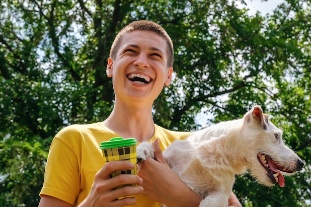 Man holds a jack russell dog and drinks coffee from a disposable cup outdoors