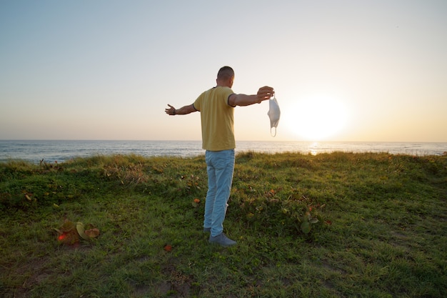 Man holds a protective face mask in his hand stands on shore of the ocean at sunrise.