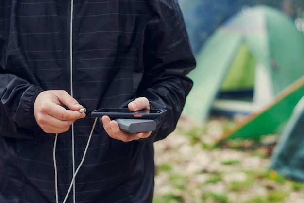 Man holds a smartphone in his hands and charges it with a power bank against the backdrop of a tourist tent in nature Portable travel charger