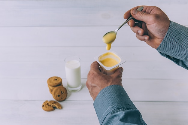 A man holds yogurt in his hand on a space of oatmeal cookies and a glass of milk.