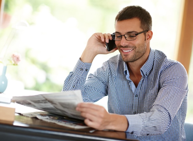 Man home and phone call while reading a newspaper morning routine and article for information Male person smile and app for conversation or networking internet and story or weekend connection