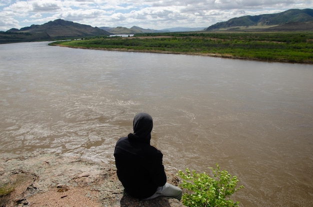 A man in a hood sits on the bank of a river in solitude. Spring, sunny weather.