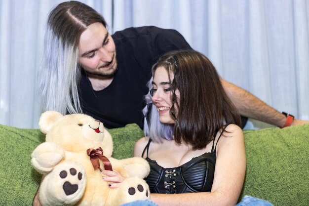 Man hugging girlfriend on sofa with teddy bear Young man giving teddy bear to girlfriend sitting on sofa