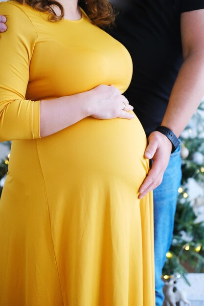 A man hugs the belly of his pregnant wife in a yellow dress Couple posing in front of the Christmas tree