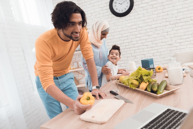 Man is cooking behind him stand his wife in hijab and son.