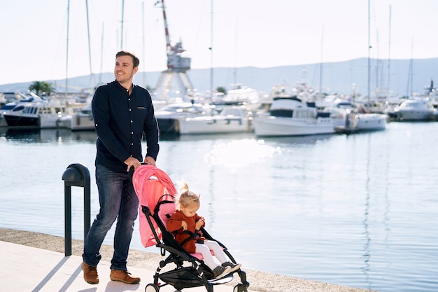 Man is pushing a pink stroller with his 2-year old daughter inside on a boat pier on a sunny day.  