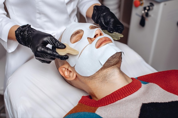 A man is on a relaxing facial massage procedure in a spa salon in a fabric mask The cosmetologist runs massagers over his face