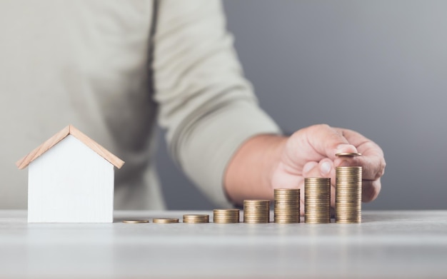 Photo man is stacking coins on a table arranging them from smallest to largest with a wooden house model beside him emphasizes the concept of saving money for future investments and financial stability