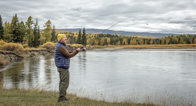 Man is standing on the river bank in the autumn forest with a fishing rod in his hands and catching fish