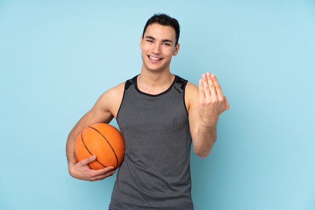 Man on isolated blue wall playing basketball and doing coming gesture