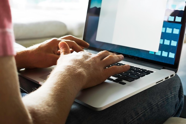 a man in jeans and a tshirt sits on the couch and prints on a laptop while working at home