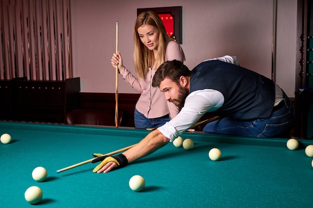 Man leaning over the table while playing snooker, he is concentrated on game, having leisure time with girlfriend