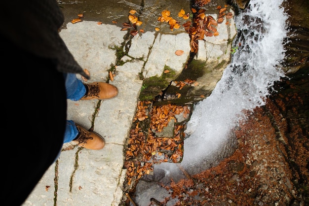 Man legs in blue jeans and brown boots sitting on the edge looking at waterfall