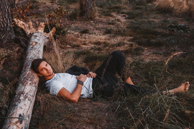 Photo man lies on the grass resting his head on a log in the middle of the forest relaxing