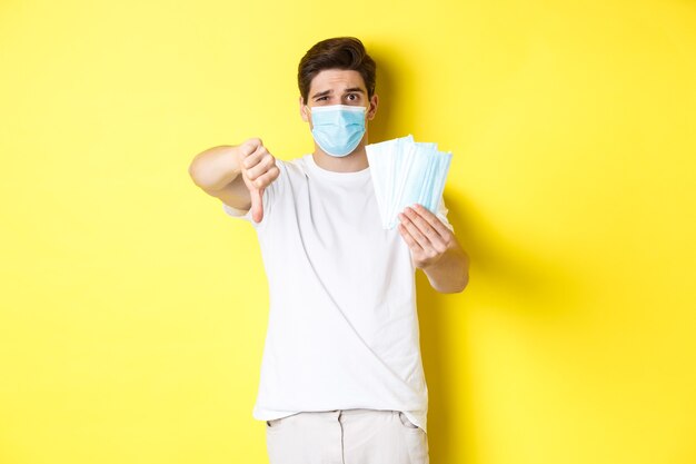 Man looking disappointed and showing thumb down, do not recommend bad medical masks, standing over yellow wall