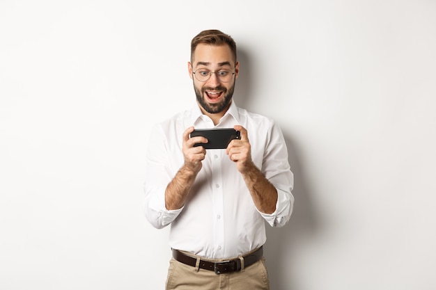 Man looking excited and surprised at mobile phone, holding smartphone horizontally, standing over white background