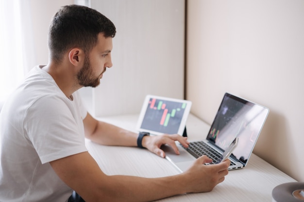 Man looking at finance chart on laptop and tablet day trader investing from home young man monitors