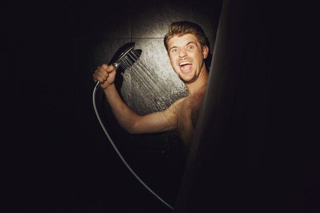 Photo man looks funny holding shower over him in black bathroom