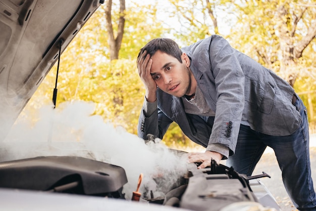 A man looks under the open hood of a car The car broke down on the road The engine is smoking