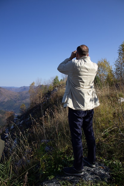 A man looks through field glasses standing on a mountain peak.