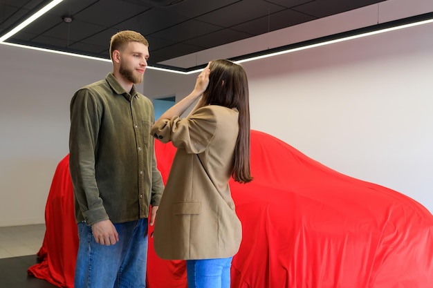 A man made a gift to a woman by buying a car at a dealership that is covered with a red cloth A young woman covers her eyes with her hands