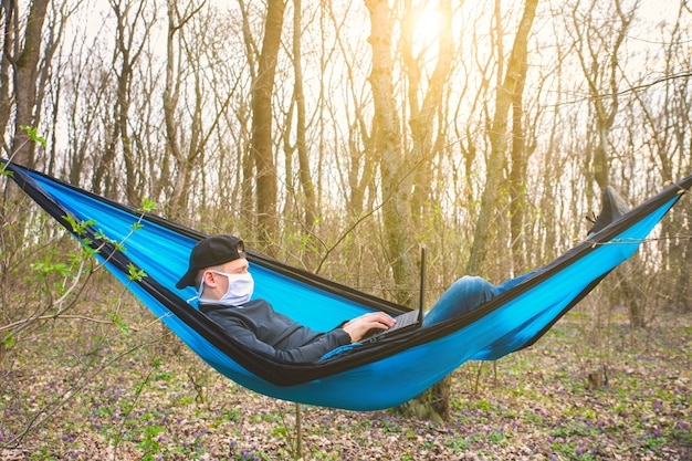 Man in mask is working on laptop in hammock. Freelancer during quarantine in the nature.  Remote work because of corona virus. No problem of unemployment for online business. Covid-19 period.