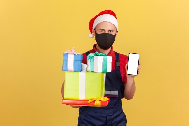 Man in mask, uniform and santa claus hat showing christmas presents and blank display smartphone