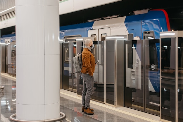 A man in a medical face mask to avoid the spread of coronavirus is holding a cellphone while waiting for a modern train on the subway. A bald guy in a surgical mask is keeping social distance.