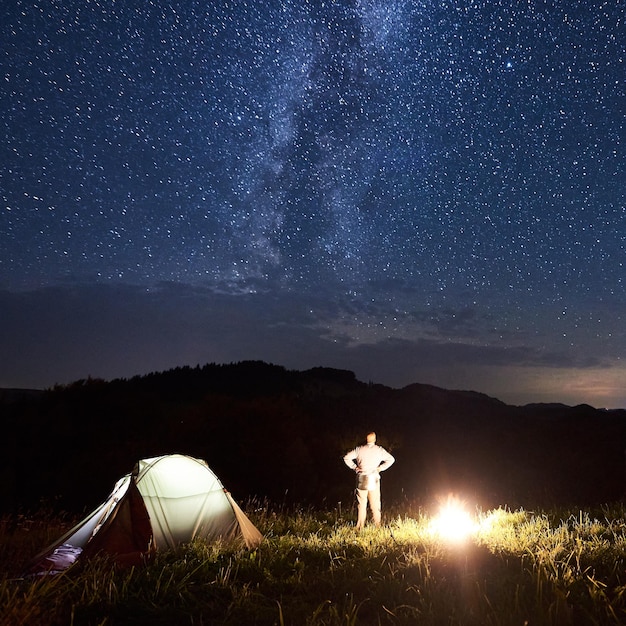 Man near tent and bonfire is meeting Milky Way at the starry sky against background of mountains