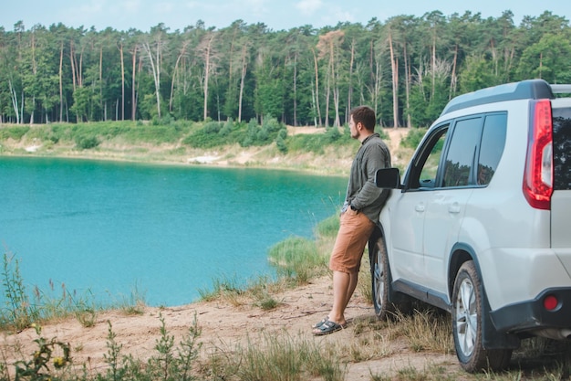 Man near white suv car at the edge looking at lake with blue water