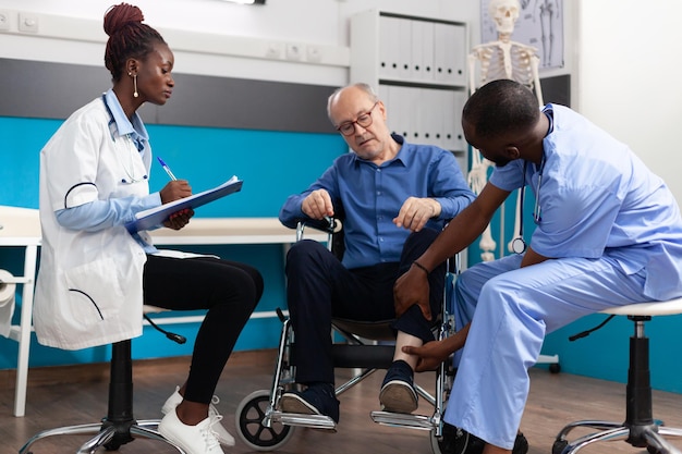 Man nurse checking injured knee leg of sick retired senior patient while therapist doctor writing medical expertise on clipboard during clinical examination in hospital office. Medicine concept