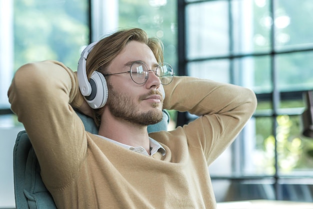 A man in an office on a chair with his arms folded is listening to music in headphones resting a