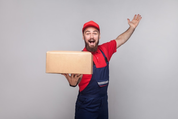 Man in overall standing holding cardboard box and toothy smiling on grey background