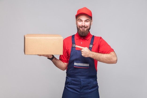 Man in overall standing holding and pointing finger to cardboard box on grey background