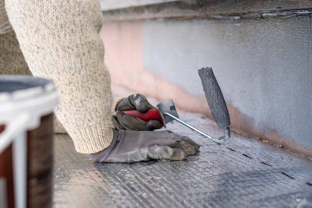 Man painting home socle with paint roller. Close up of rolling grey paint on building plinth. Home renovation