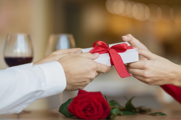 A man passes a white giftbox with a red ribbon to his girlfriend. A young woman takes a present from her boyfriend. Two glasses of wine and a rose on the cafe's table. Valentine's day concept.