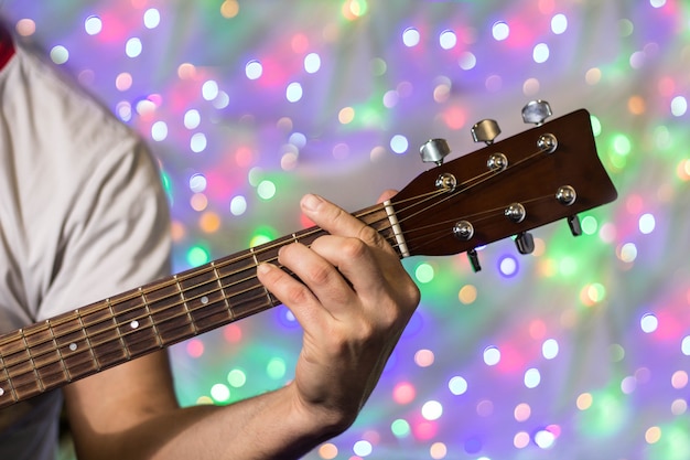 Man playing on acoustic guitar. Closeup fingers on guitar neck against christmas blurred bokeh lights on background.